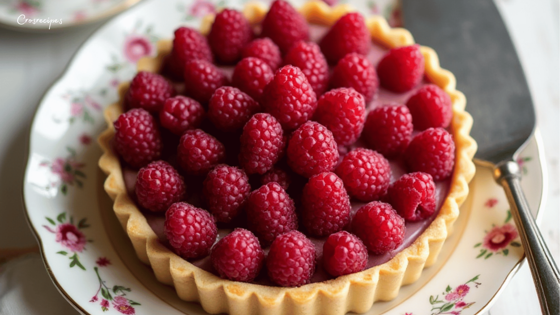 Une tarte à la framboise décorée de framboises fraîches, saupoudrée de sucre glace, servie sur une assiette élégante.