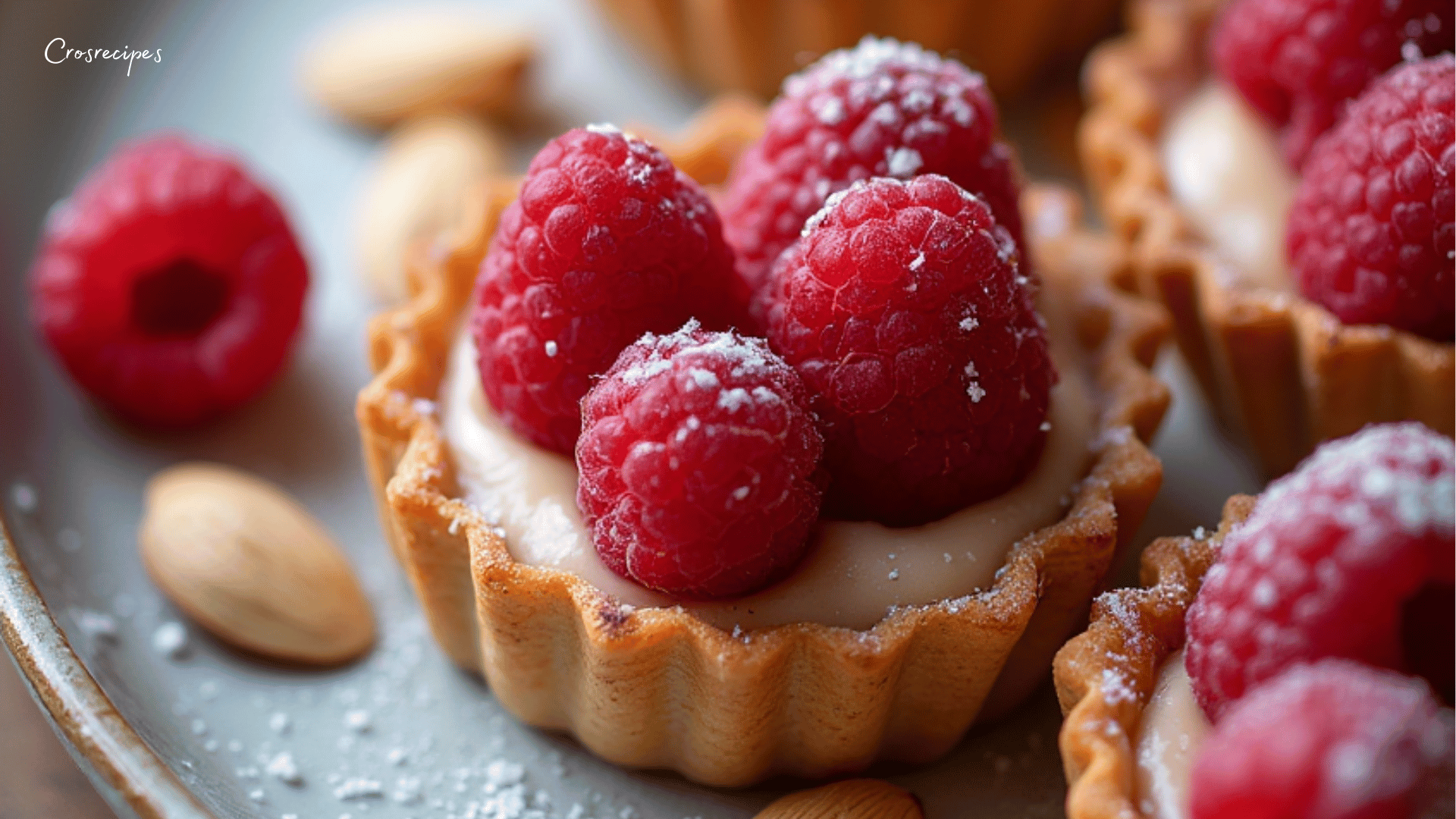 Tartelettes aux framboises et aux amandes, garnies de crème d’amande et saupoudrées de sucre glace, servies sur une assiette.