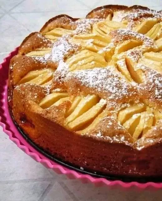 Un gâteau doré aux pommes et aux noix, saupoudré de sucre glace, avec des morceaux de pommes visibles sur le dessus.