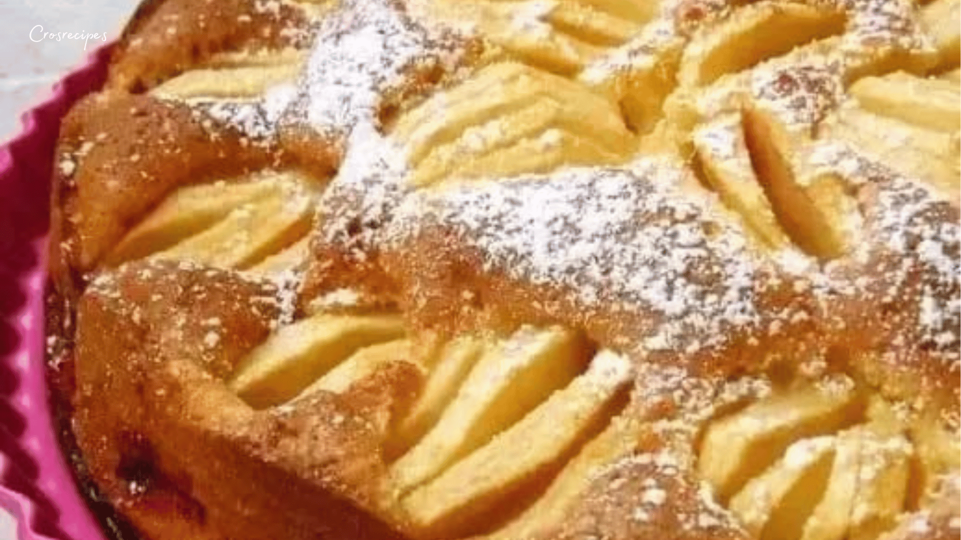 Un gâteau doré aux pommes et aux noix, saupoudré de sucre glace, avec des morceaux de pommes visibles sur le dessus.