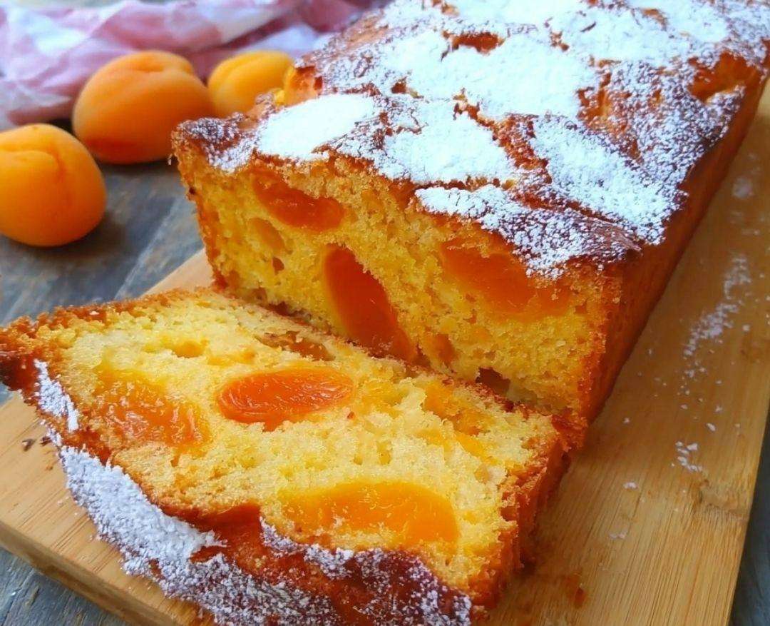 Tranche de cake au yaourt aux abricots, moelleux et doré, parsemé d’amandes effilées, servie sur une assiette avec des abricots frais.