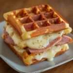 Assiette de gaufres salées dorées garnies de jambon et fromage fondant, servies en croque-gaufres croustillants avec salade verte sur le côté.