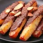 Orangettes au chocolat noir brillantes décorées d’éclats d’amandes grillées, alignées sur une feuille de papier cuisson.