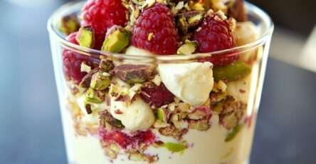 Verrines gourmandes à la crème mascarpone vanillée, framboises fraîches et pistaches concassées, présentées en couches colorées dans des verres transparents.