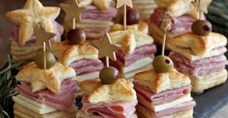 Mini sandwichs étoiles feuilletées garnies de jambon et fromage, montées en sandwichs et décorées d’une olive sur un pic, présentées sur un plateau festif