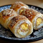 Roulés de pain garnis à l’œuf, dorés au four et roulés dans des graines de sésame, présentés en tranches montrant l’œuf dur au centre