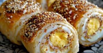 Roulés de pain garnis à l’œuf, dorés au four et roulés dans des graines de sésame, présentés en tranches montrant l’œuf dur au centre