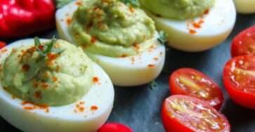 Demi-œufs mimosa garnis d’une crème verte à l’avocat, décorés de tomates cerises, herbes fraîches et paprika, joliment présentés sur un plat de service