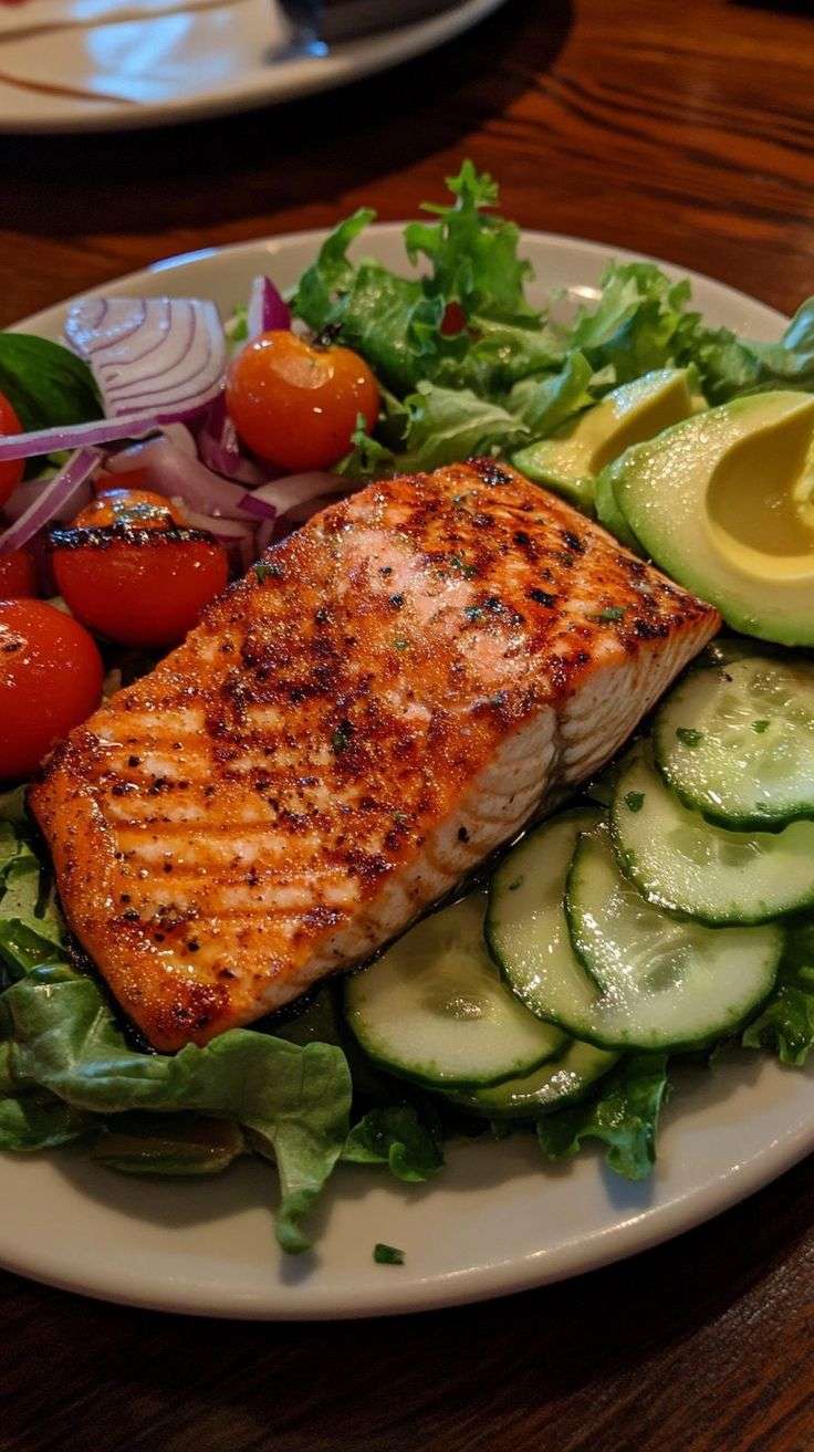 Salade de saumon grillé doré, tranches d’avocat mûr, concombre croquant, tomates cerises et vinaigrette citronnée dans une assiette blanche.