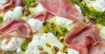 Pizza dorée au four garnie de pesto vert, burrata fondante, tranches fines de prosciutto et pistaches concassées, arrosée d’huile d’olive et décorée de basilic frais.