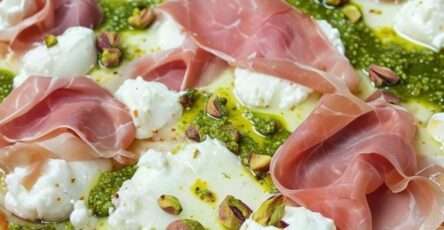 Pizza dorée au four garnie de pesto vert, burrata fondante, tranches fines de prosciutto et pistaches concassées, arrosée d’huile d’olive et décorée de basilic frais.