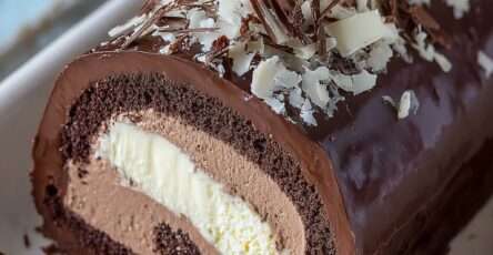 Bûche roulée au chocolat nappée d’un glaçage brillant, garnie d’un cœur vanille visible à la coupe et décorée de copeaux de chocolat noir et blanc