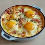 Œufs cocotte au fromage et lardons servis en ramequins, blancs pris et jaunes coulants, accompagnés de pain grillé.