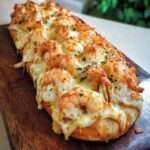 Baguette gratinée coupée en deux, garnie de crevettes, sauce crémeuse à l’ail et fromage fondu doré, parsemée d’herbes fraîches.