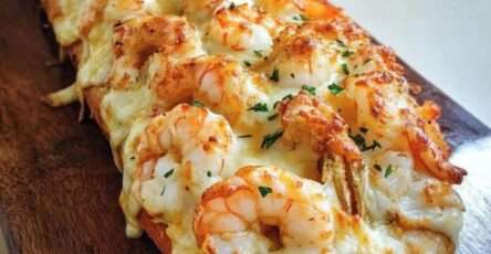 Baguette gratinée coupée en deux, garnie de crevettes, sauce crémeuse à l’ail et fromage fondu doré, parsemée d’herbes fraîches.