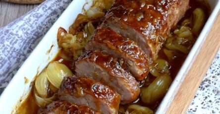 Rôti de porc au miel et aux oignons fondants servi en tranches avec sauce brillante, oignons caramélisés et herbes, présenté dans un plat rustique.
