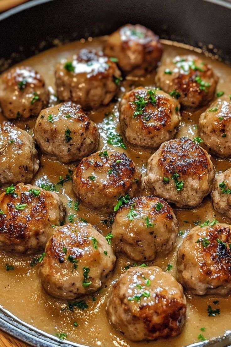 Boulettes de viande dorées mijotées dans une sauce brune onctueuse avec persil frais, servies dans une poêle rustique.