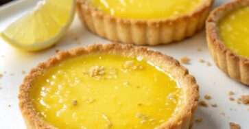 Tartelettes au citron garnies d’une crème lisse et brillante, décorées de zestes de citron frais, présentées sur une assiette élégante.