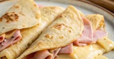 Crêpes salées au jambon et au fromage servies chaudes dans une assiette, garnies de fromage fondant, pliées en triangle, avec une texture dorée et moelleuse.