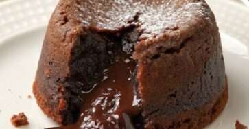 Fondant au chocolat individuel, légèrement craquelé à l’extérieur, avec un cœur coulant intense au chocolat noir, servi chaud sur une assiette avec une texture brillante.