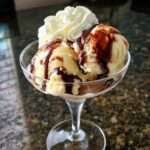 Coupe glacée vanille chocolat servie dans un verre à dessert, avec boules de glace vanille, sauce chocolat tiède, chantilly, copeaux de chocolat et amandes effilées.