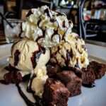 Brownie glacé servi en coupe avec morceaux de brownie fondant, boules de glace vanille, sauce chocolat tiède, chantilly et copeaux de chocolat.
