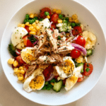 Macédoine revisitée au thon et œufs servie dans un grand plat, avec pommes de terre, tomates cerises, concombre, brocoli, maïs et sauce crémeuse.