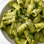 Pâtes crémeuses au pesto vert servies dans une assiette creuse avec parmesan râpé, basilic frais et sauce verte onctueuse.