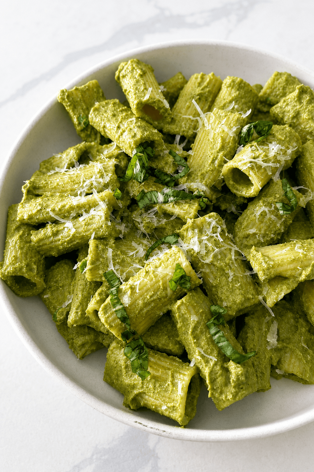 Pâtes crémeuses au pesto vert servies dans une assiette creuse avec parmesan râpé, basilic frais et sauce verte onctueuse.