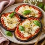 Rondelles d’aubergines façon mini-pizzas garnies de sauce tomate, de mozzarella fondue et d’origan, dorées au four sur une plaque de cuisson.