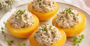 Demi-pêches jaunes garnies d’une préparation crémeuse au thon et à la mayonnaise, décorées de ciboulette, de persil frais et de poivre, servies froides sur un lit de salade verte.