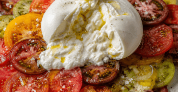 Burrata crémeuse déposée au centre d’un carpaccio de tomates anciennes rouges, jaunes, vertes et orangées, assaisonné d’huile d’olive, de basilic frais et de poivre noir.