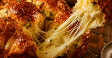 Brioche pull-apart dorée composée de boules moelleuses serrées les unes contre les autres, dont une portion est détachée pour révéler un cœur de Comté fondu et filant.