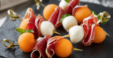 Mini-brochettes apéritives composées de billes de melon orange, de jambon de Bayonne plié et de mozzarella, décorées de basilic frais.