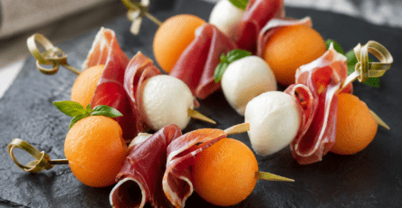 Mini-brochettes apéritives composées de billes de melon orange, de jambon de Bayonne plié et de mozzarella, décorées de basilic frais.