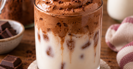 Verre de lait chocolat fouetté servi avec des glaçons, surmonté d’une mousse aérienne au cacao et au sucre, pour une boisson fraîche et crémeuse.
